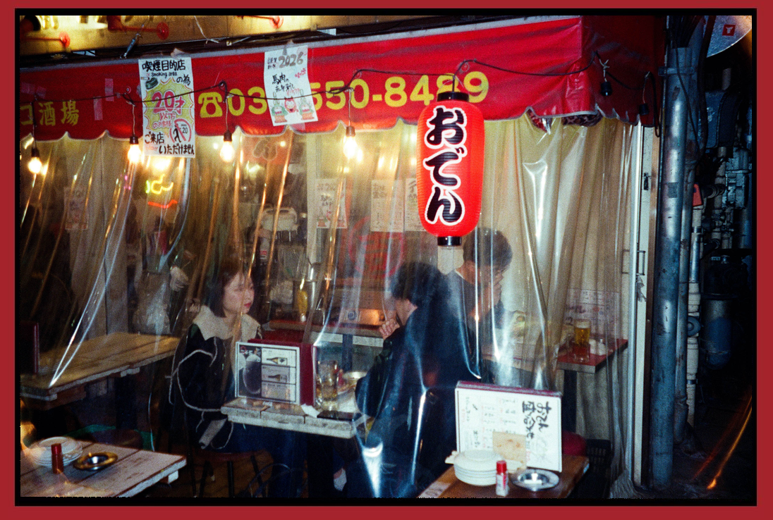 People dining inside a Japanese restaurant with plastic curtains, illuminated by warm string lights, red signage, and a red paper lantern.