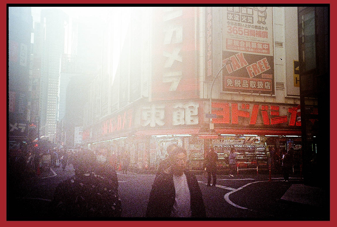 A city street scene in Japan with neon signs and advertisements in Japanese, pedestrians walking, and sunlight creating a hazy atmosphere.