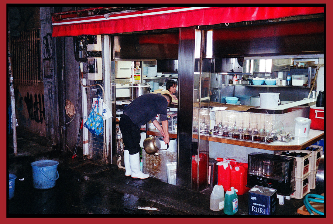 A person is washing a pot outside a small kitchen or food stall, with various kitchen items and supplies visible inside.