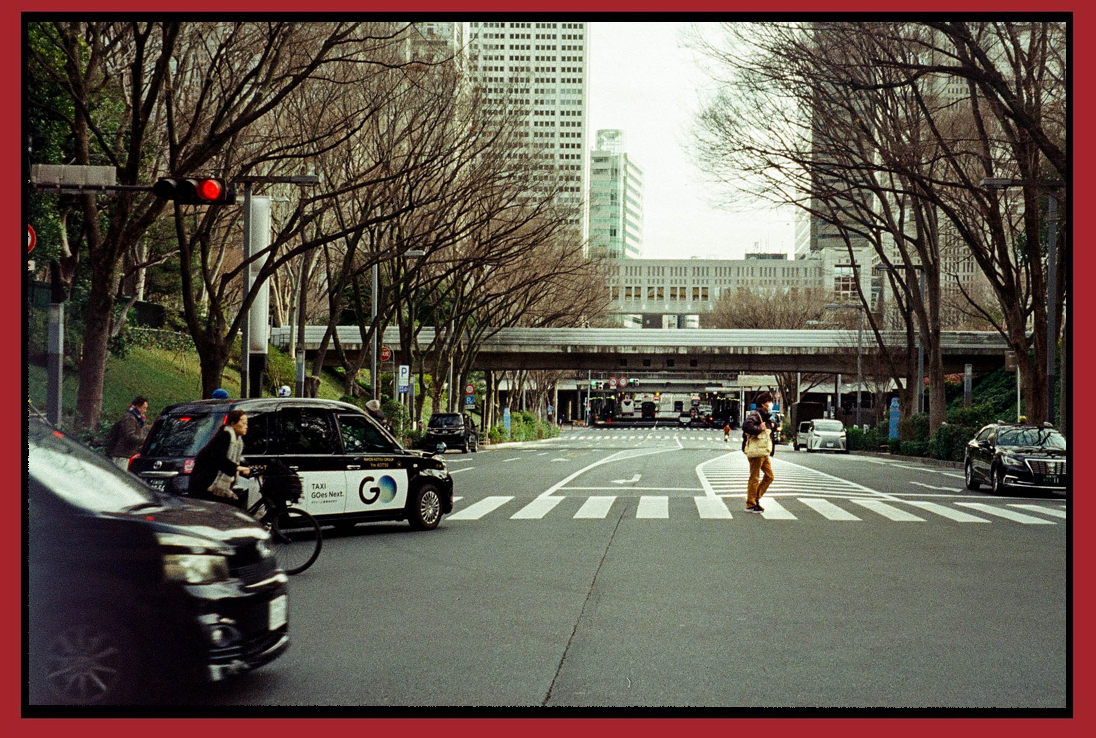 City street intersection with pedestrians, cars, and a crosswalk, with leafless trees and tall buildings in the background.