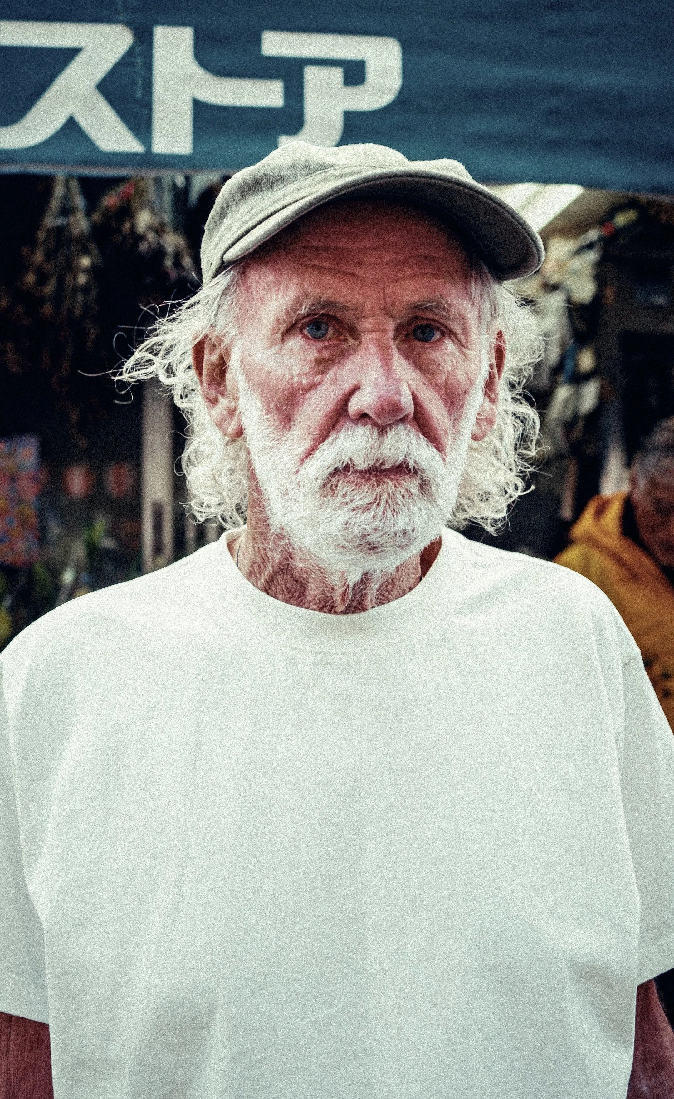 An elderly man with a white beard and mustache wearing a light-colored cap and a white T-shirt, standing outdoors in front of a marketplace background.