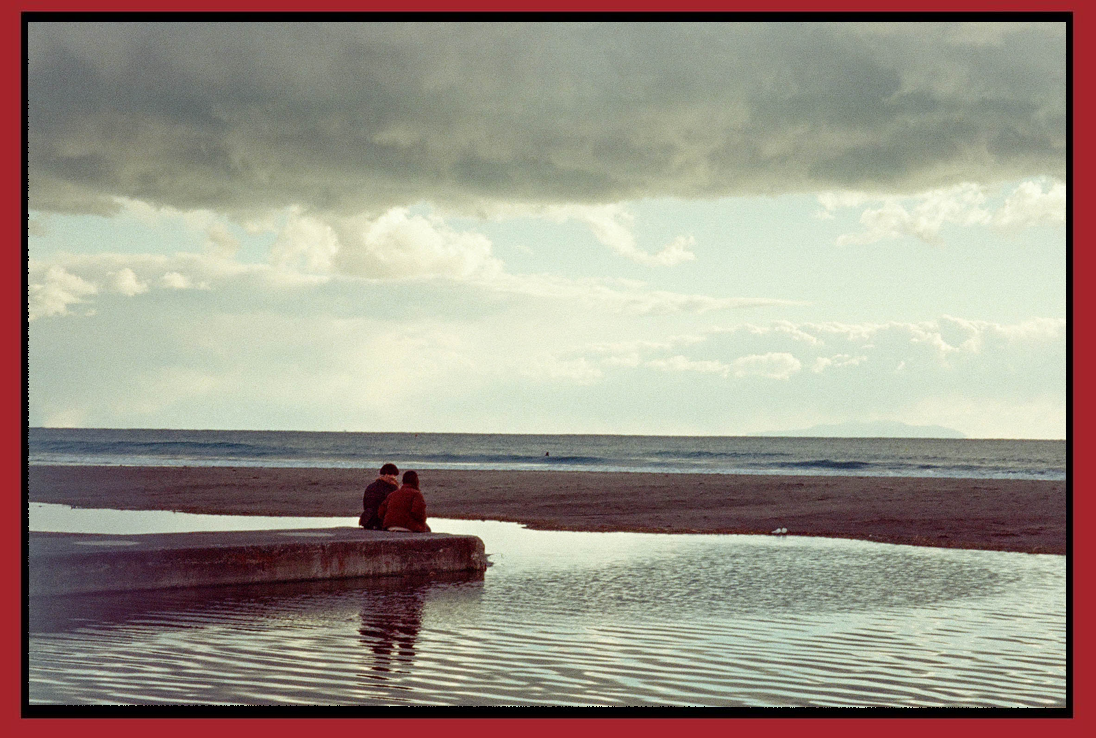 Two people sitting on a concrete ledge near a body of water on a beach, with overcast skies and the ocean in the background.