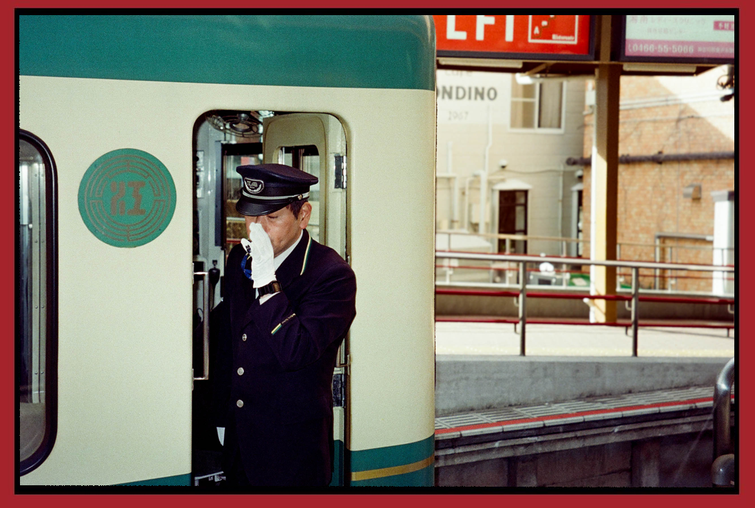 A train conductor in a navy uniform and cap, wearing white gloves, is talking into a radio or phone while standing next to a train at a station.