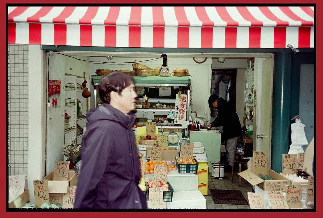 A small grocery shop with produce like oranges and vegetables, with handwritten signs in Japanese, under a red and white striped awning, and a man shopping inside.