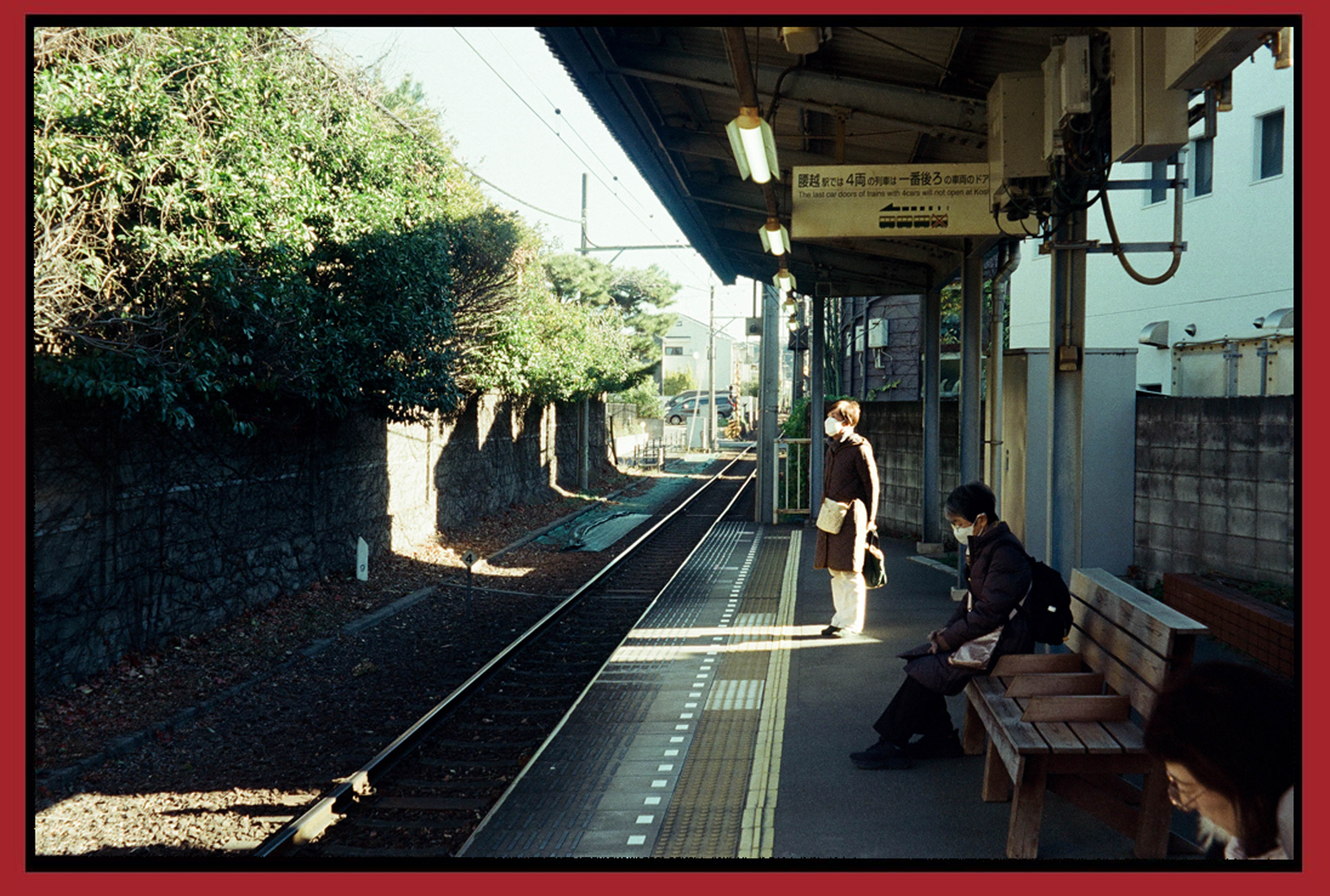 People waiting on a train platform, two women sitting on a bench and one woman standing, all wearing face masks, with train tracks and greenery in the background.
