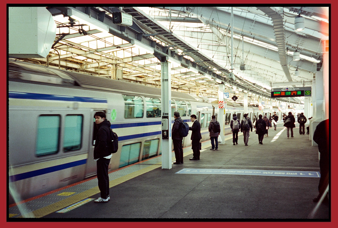 People boarding and standing on a platform at a busy train station with trains and electronic departure boards overhead.