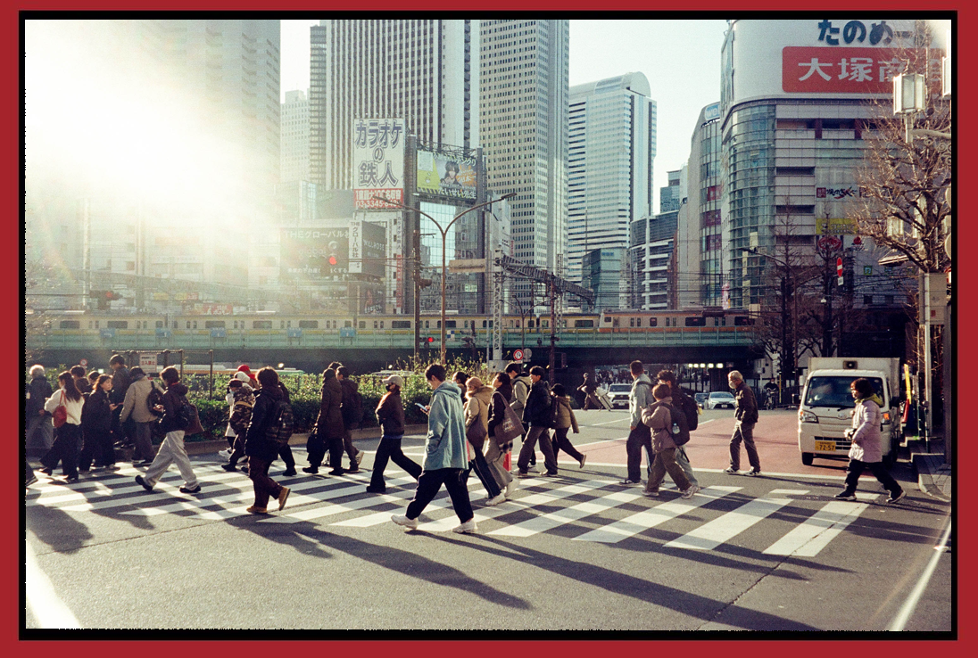 People crossing a busy city street in front of tall skyscrapers, with sunlight and a train passing overhead.