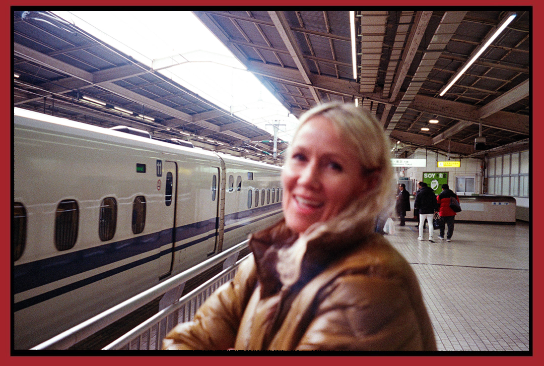 A smiling woman standing on a train station platform with a high-speed train in the background.