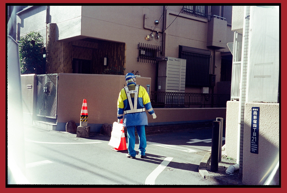 A construction worker in blue and yellow uniform with a blue helmet standing on a street, holding an orange traffic cone and a white bag, near several orange traffic cones around a corner in a city neighborhood.