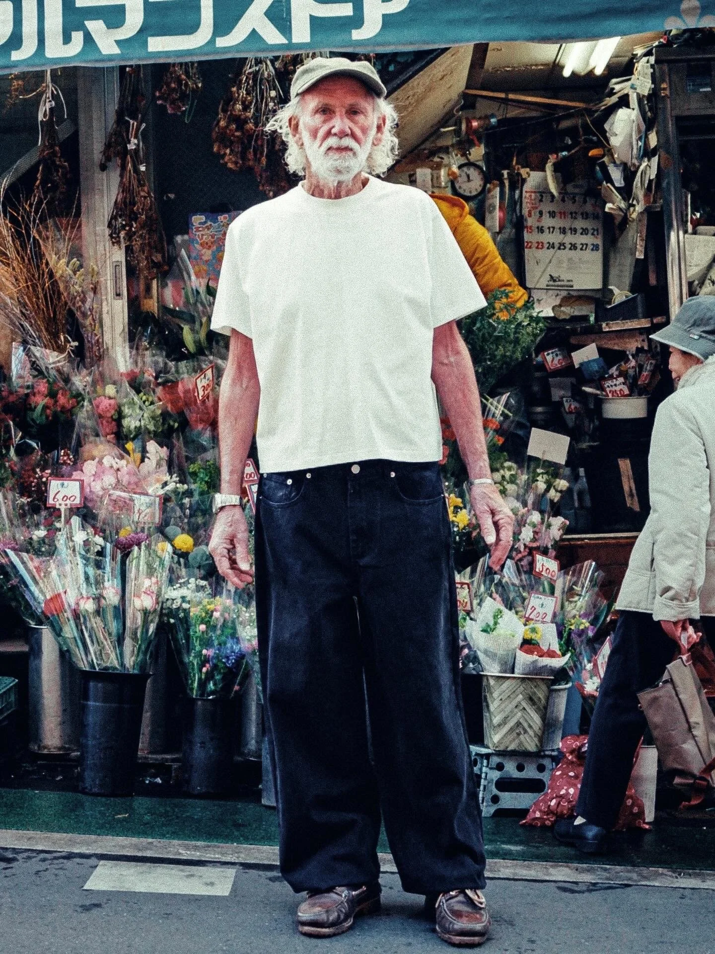 An older man with white hair and a beard standing in front of a florist shop. The shop displays colorful flowers wrapped in plastic and some signs with prices. He is wearing a white t-shirt, dark pants, and a cap.