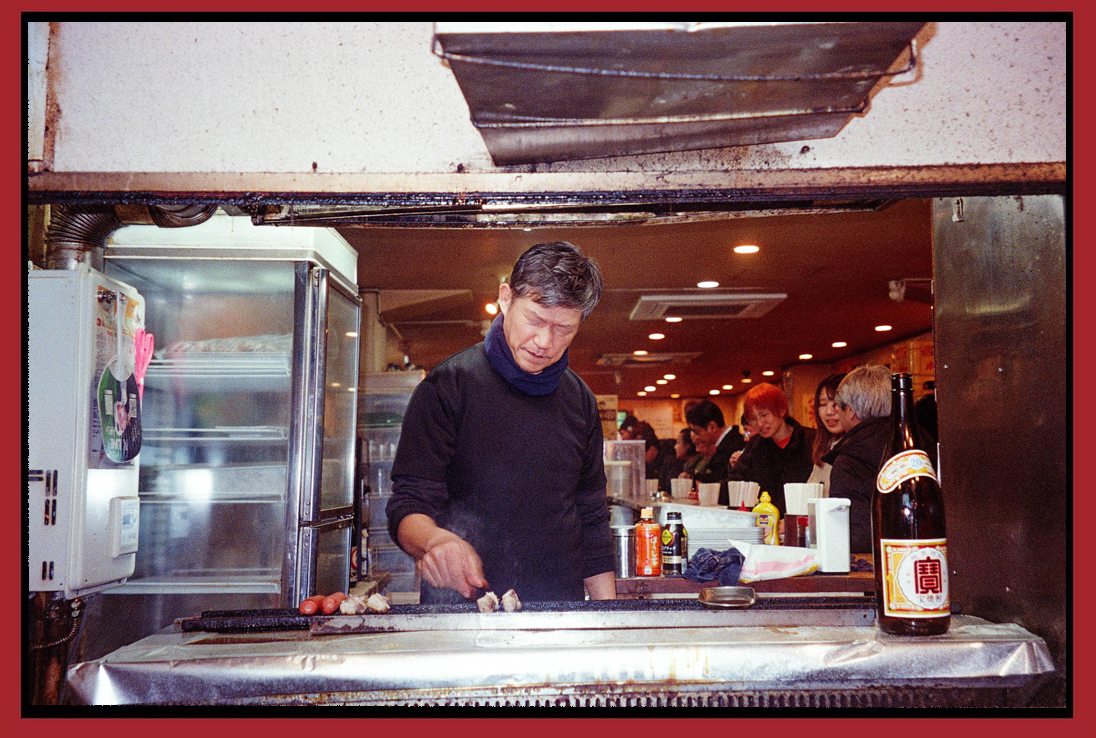 A man cooking meat on a grill in a busy restaurant kitchen with customers in the background.