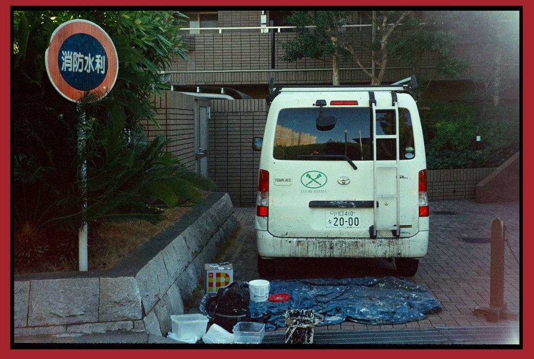 A white work van parked on a brick driveway next to a raised flower bed with green foliage. A sign with Japanese characters is on the left. In front of the van, there are painting supplies, including a paint bucket, paint cans, brushes, and a tarp.