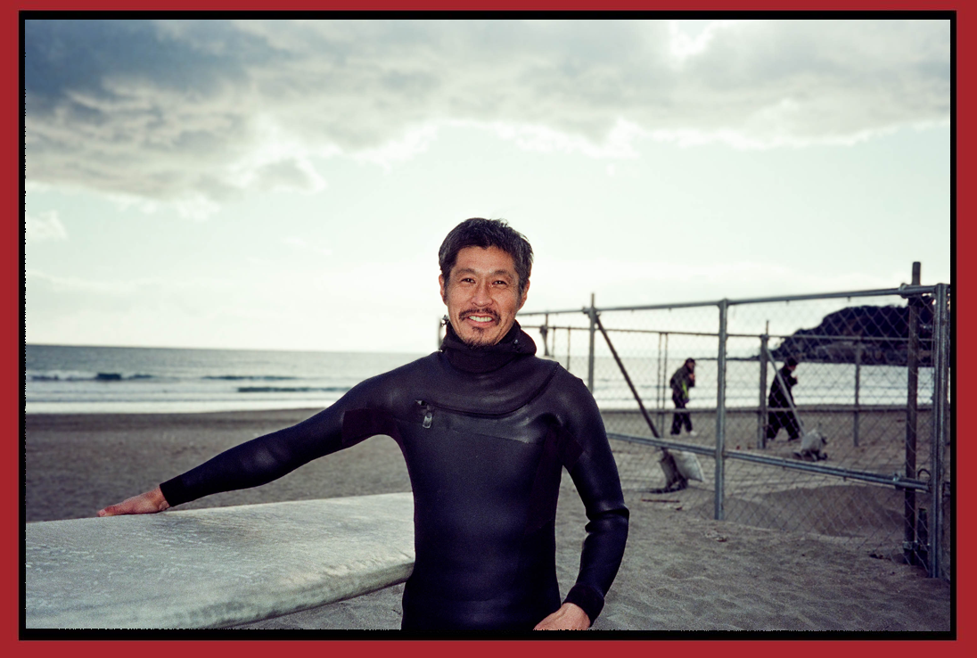 Man in a black wetsuit standing on a beach with a surfboard, holding it with his right hand, smiling at the camera, with the ocean and cloudy sky in the background.