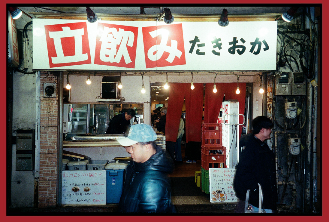 Exterior of a Japanese restaurant with a white signboard displaying red and black Japanese characters. Inside, there are some people and kitchen staff preparing food, with a red curtain dividing the space.