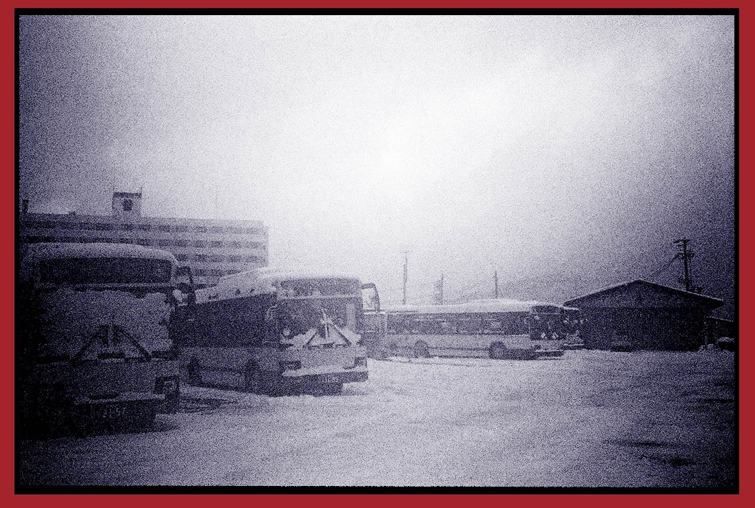 A snow-covered lot with several parked buses, a small building, and in the background, a multi-story building and utility poles, under overcast skies.
