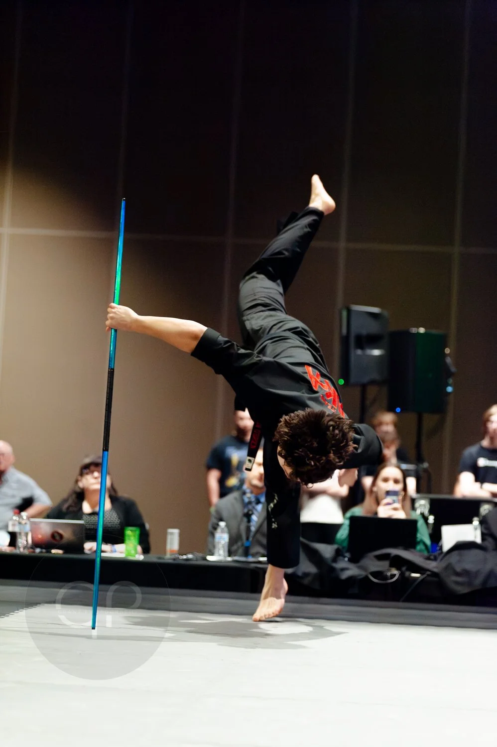 A person performing a breakdancing move on a stage, balancing on one hand with legs raised, holding a staff, while an audience watches in the background.