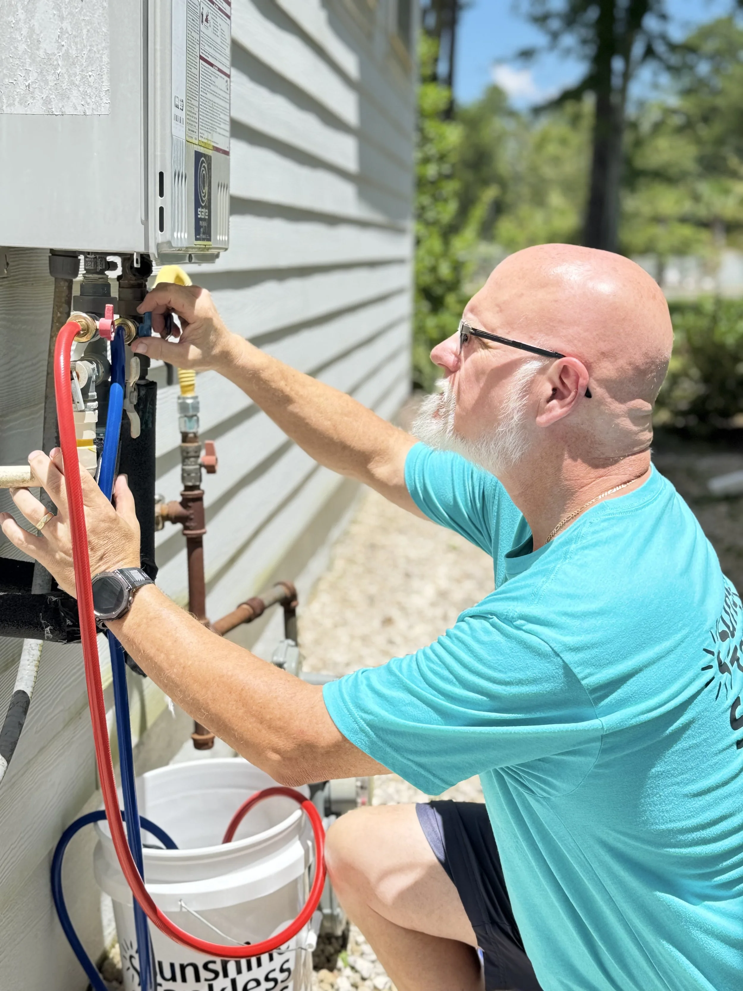A man with a bald head, glasses, and a white beard working on an outdoor gas or water appliance, adjusting valves or connections. He's wearing a turquoise T-shirt and black shorts, and a bucket with hoses is nearby.