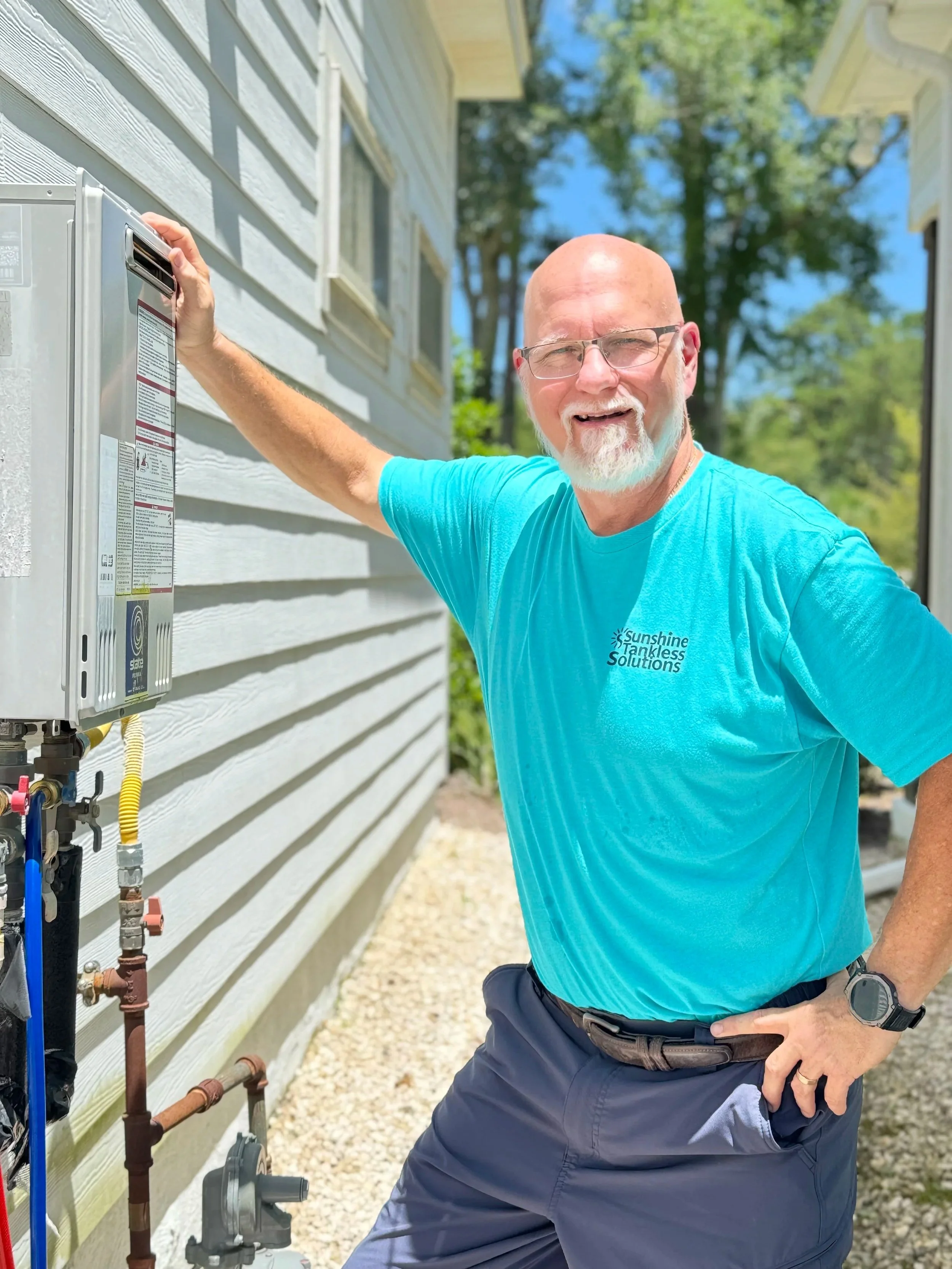 A man with a beard and glasses, wearing a blue t-shirt with a company logo, smiling and standing outdoors next to an outdoor water heater or boiler attached to a house wall.