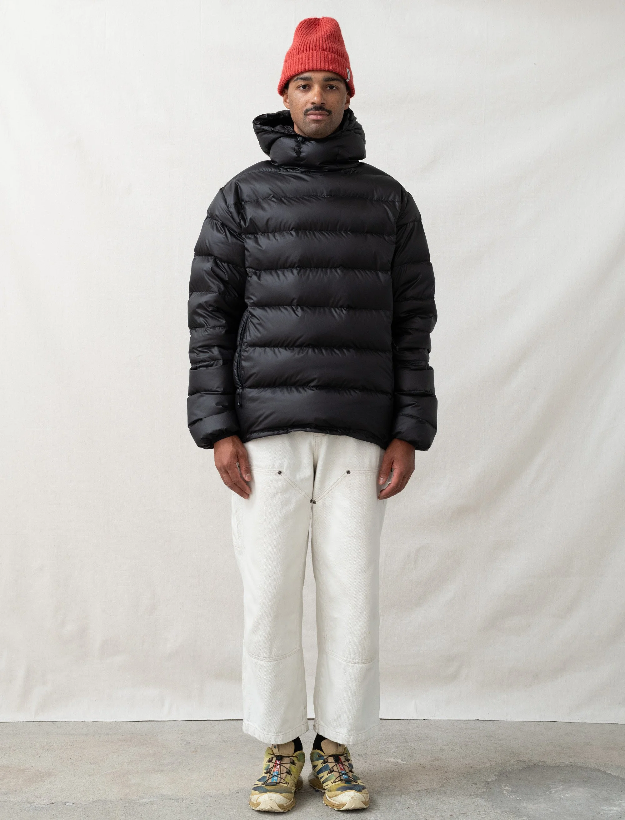 Man wearing a red knit beanie, black puffer jacket, white wide-leg pants, and hiking shoes, standing against a plain white background.