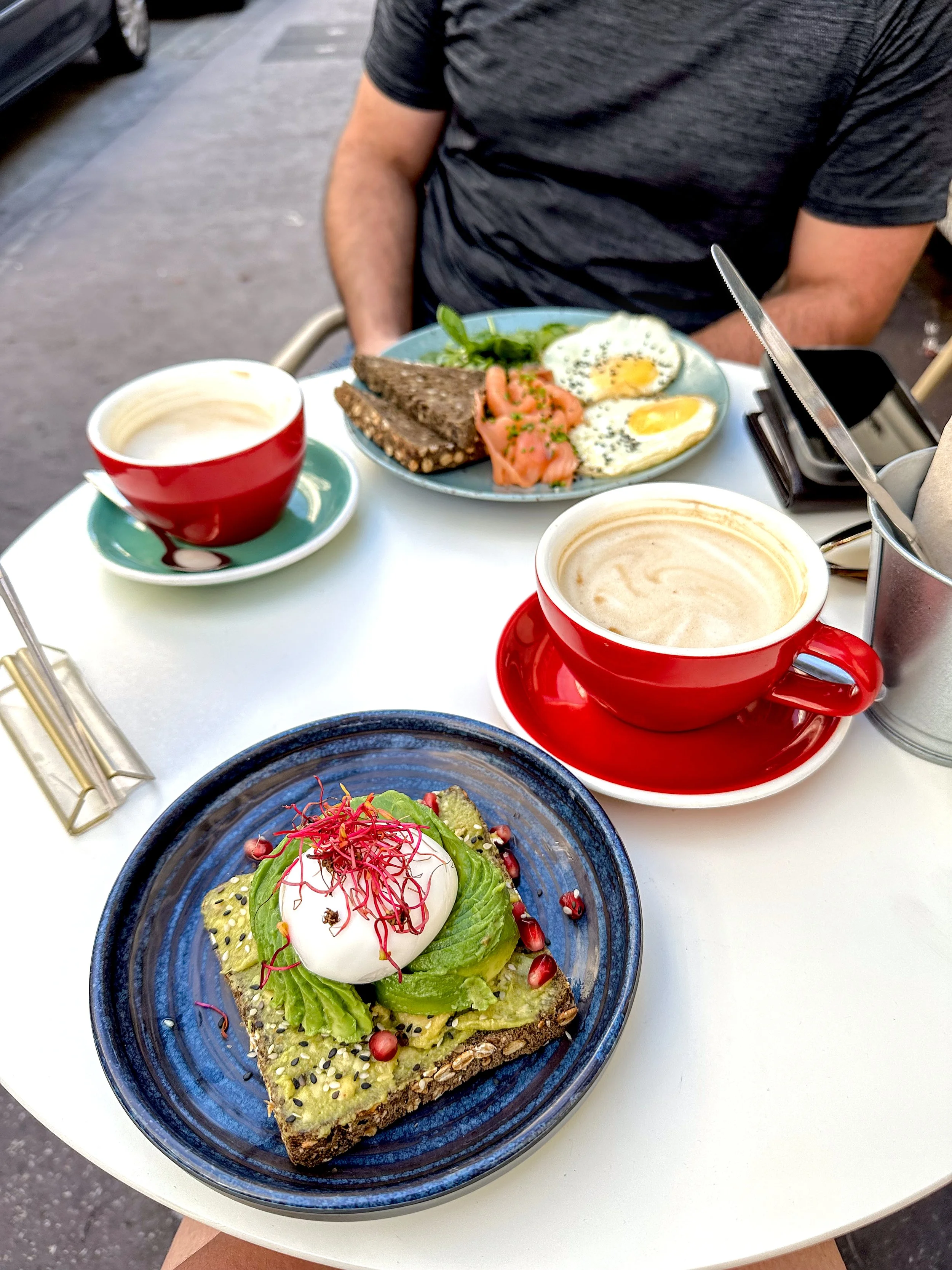 A cafe table with two red cups of coffee and plates of food, including avocado toast with a poached egg and a plate with smoked salmon and fried eggs
