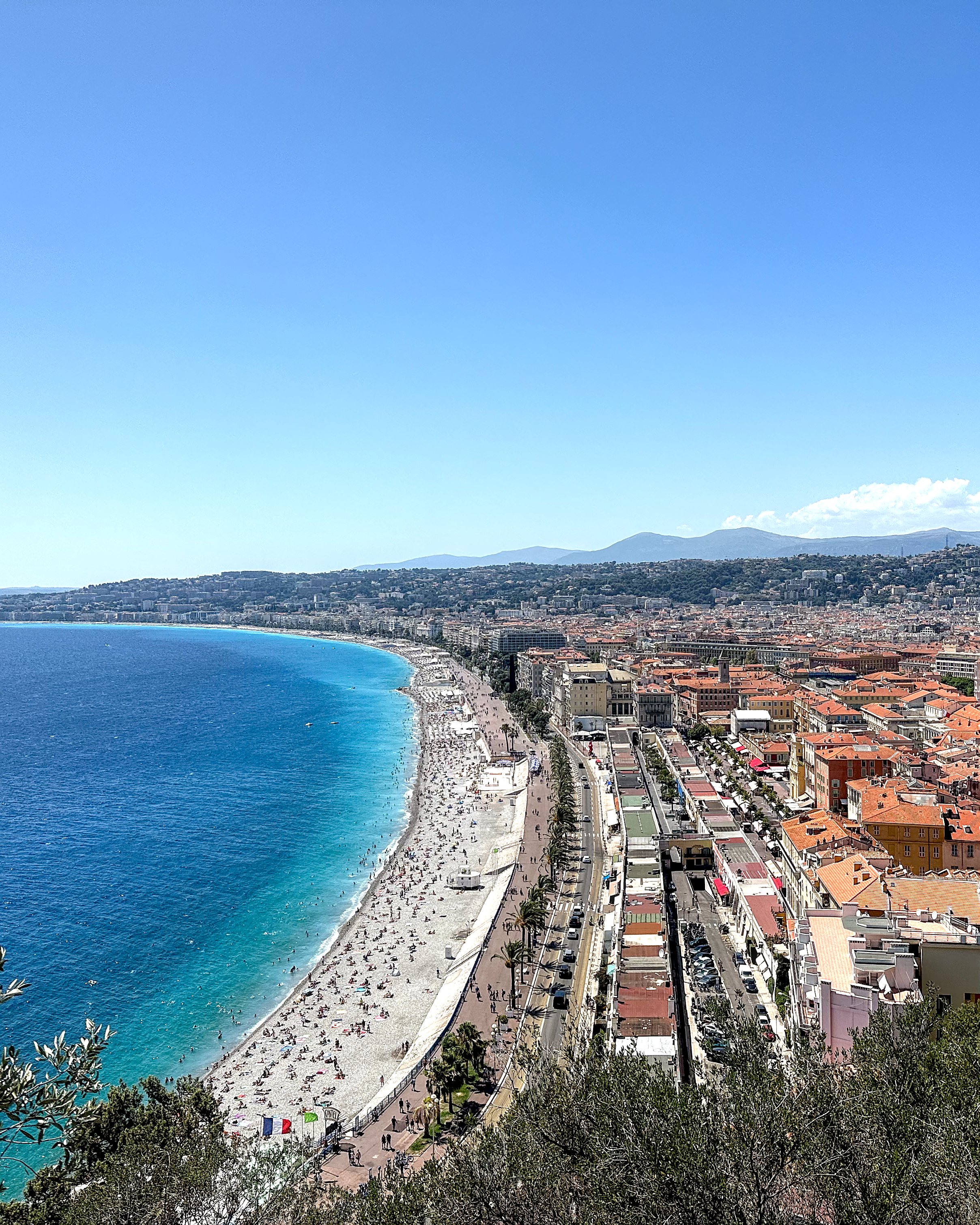 An elevated view of the Promenade des Anglais in Nice, showing the pebble beach, the blue sea, and the city with its red rooftops