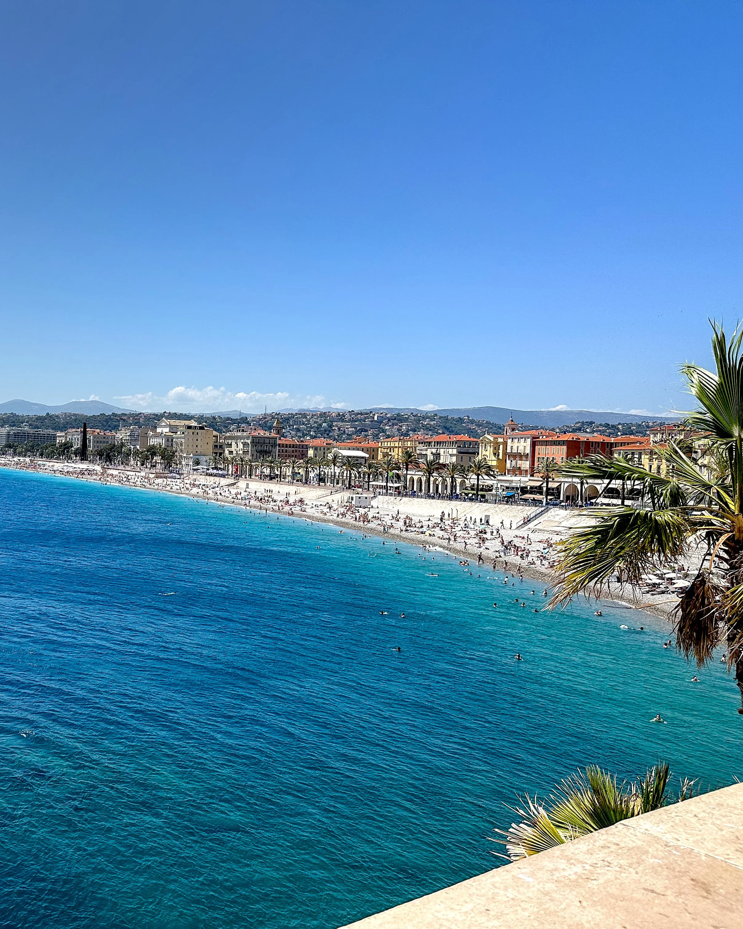 A panoramic view of the pebble beach in Nice, with people sunbathing and swimming in the bright blue Mediterranean Sea