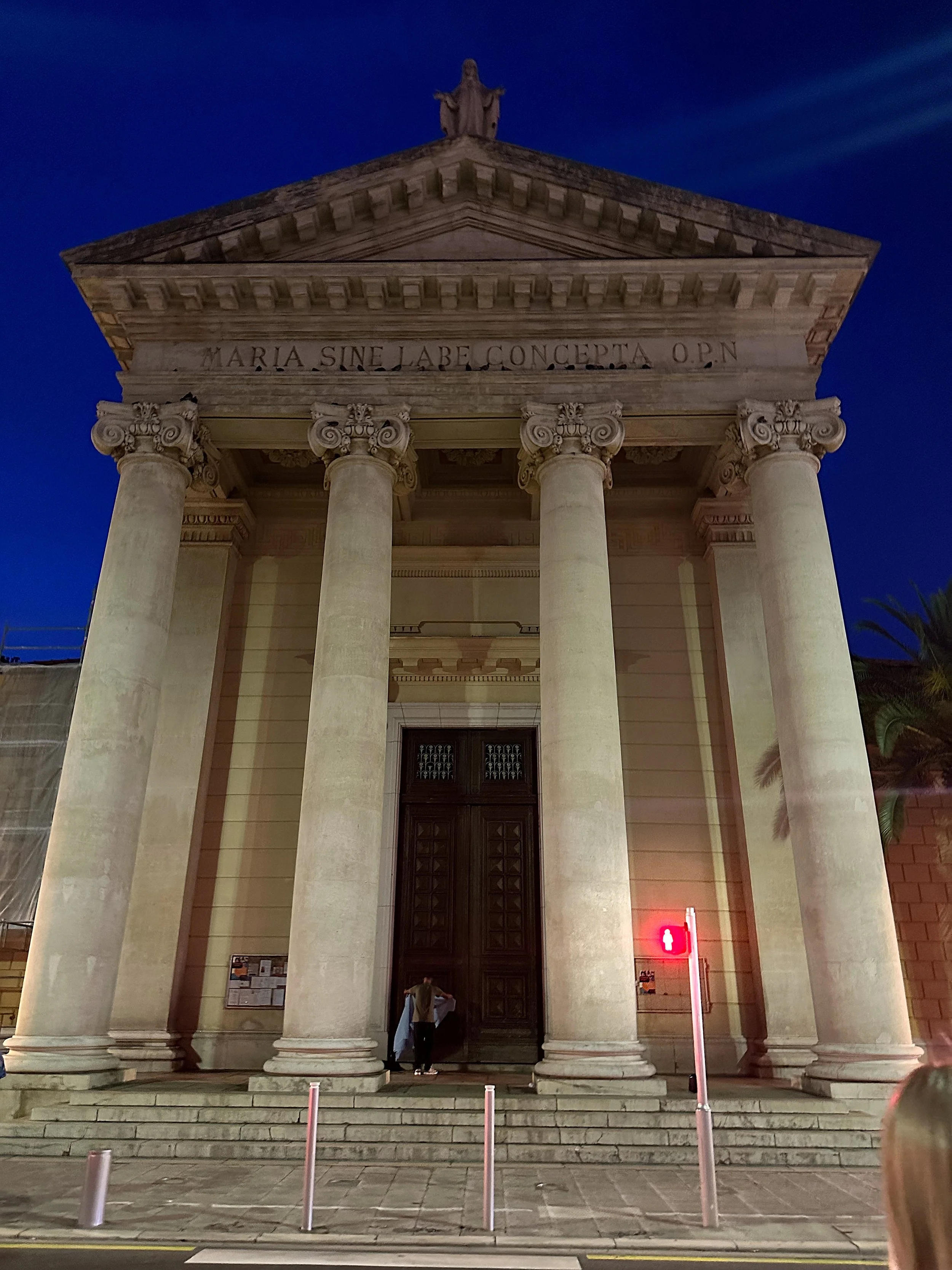 The lit-up exterior of the Église Notre-Dame-du-Port in Nice at night, showing its neoclassical columns and a person sitting on the front steps