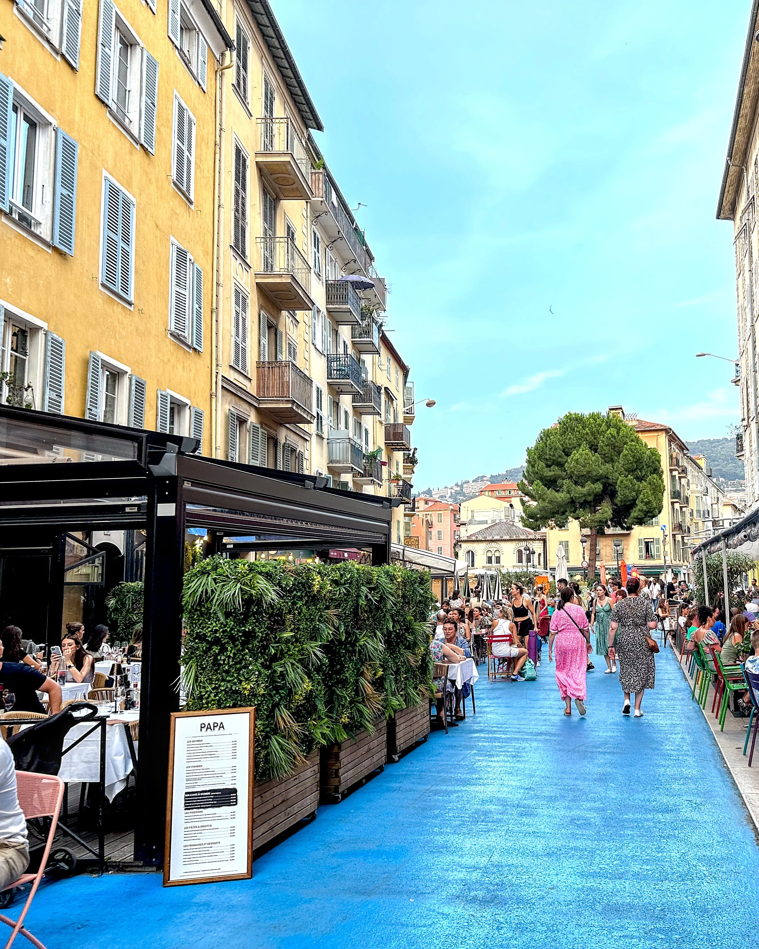A narrow street in Nice with a bright blue painted road, lined with restaurants and people eating at outdoor tables