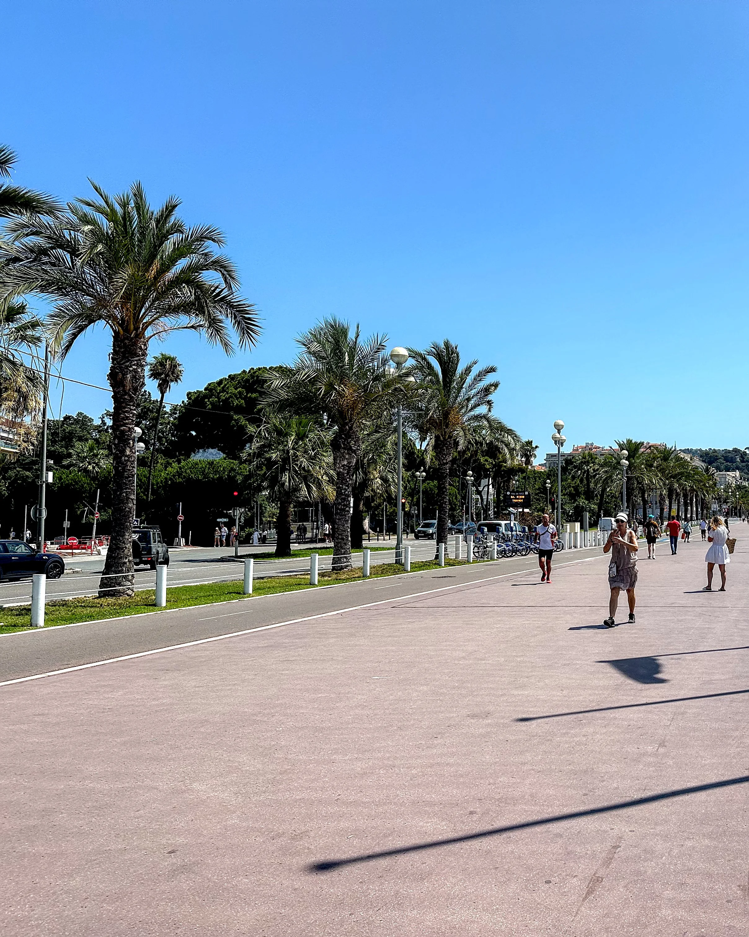 A view of the Promenade des Anglais in Nice, showing a wide walkway lined with palm trees and a few people walking