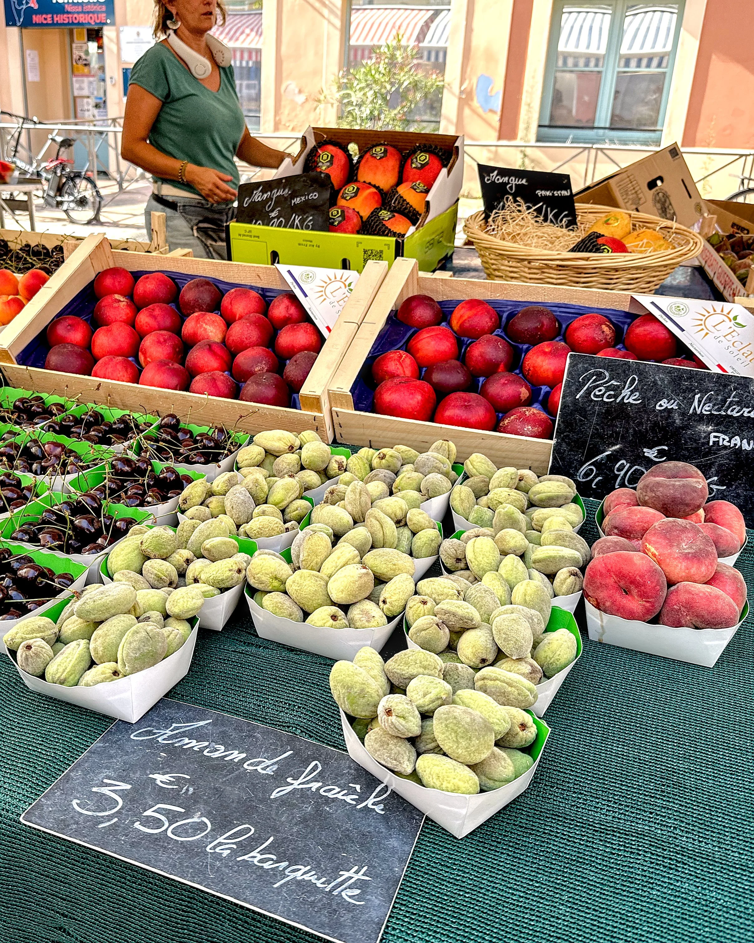 A close-up of a fruit stall in Nice, showing baskets of fresh green almonds, red peaches, and cherries