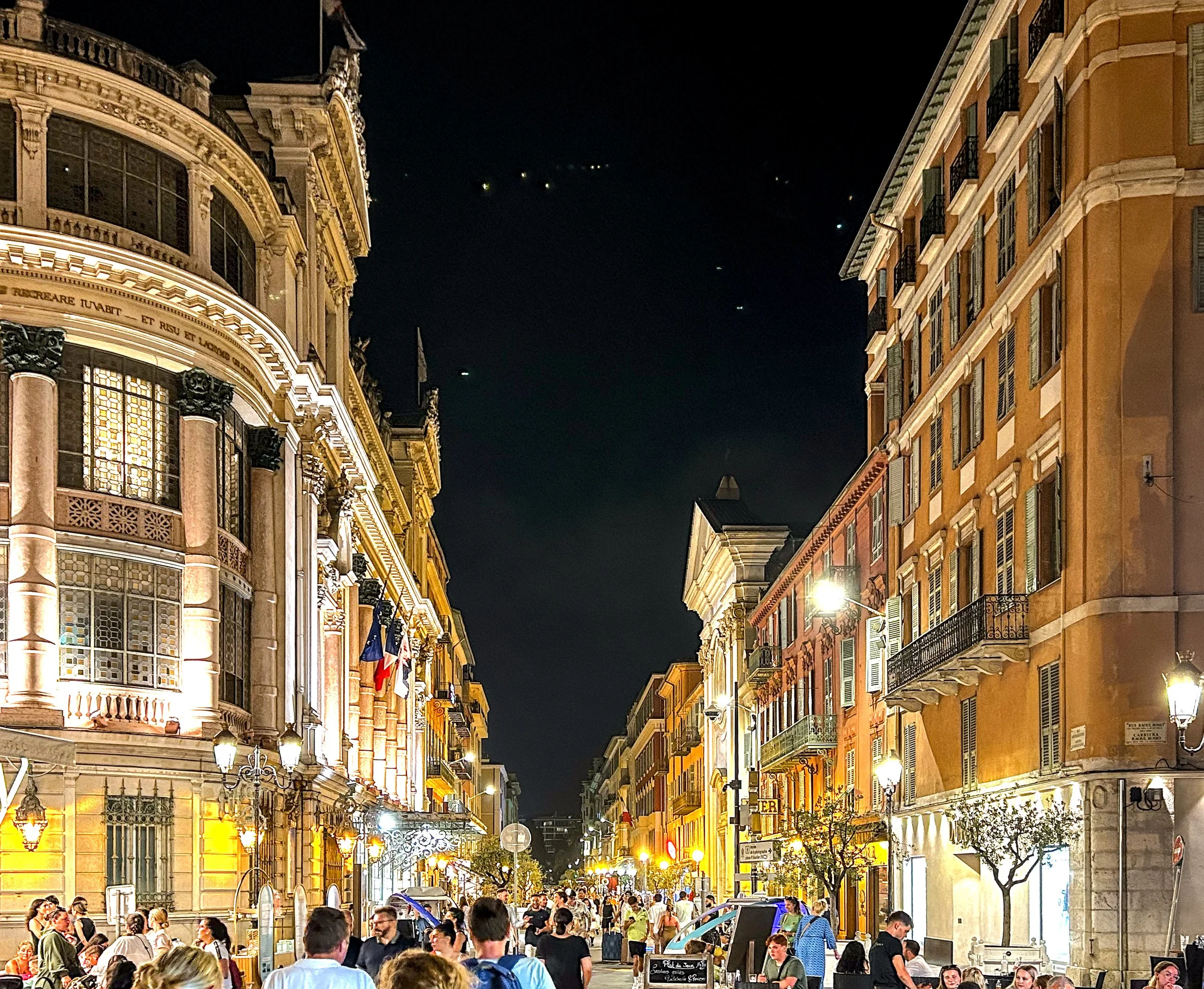 A narrow street in Nice at night, with people dining at outdoor restaurants and the buildings on both sides illuminated