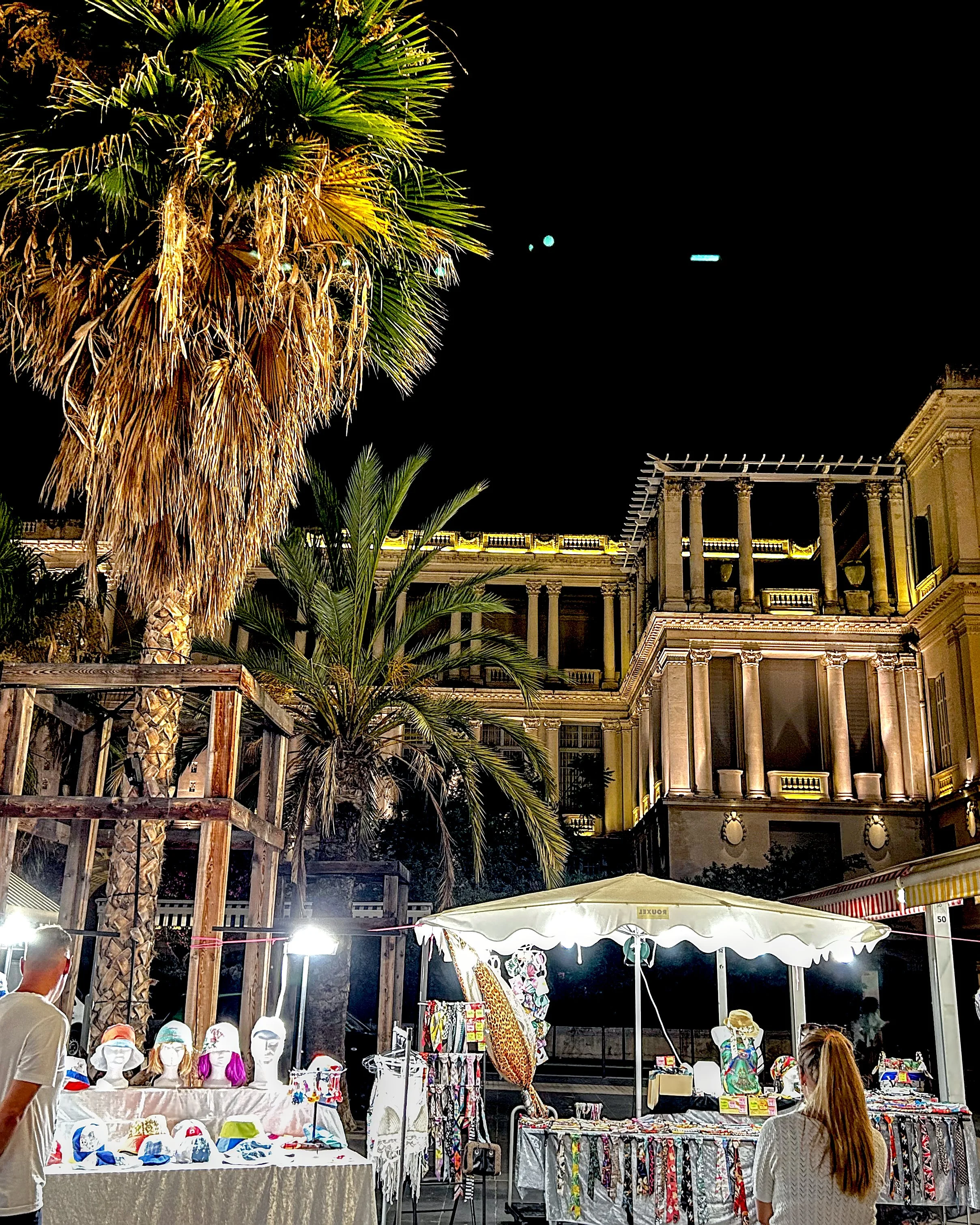 A night market in Nice, with a stall selling hats and scarves in front of a palm tree and a lit-up historic building