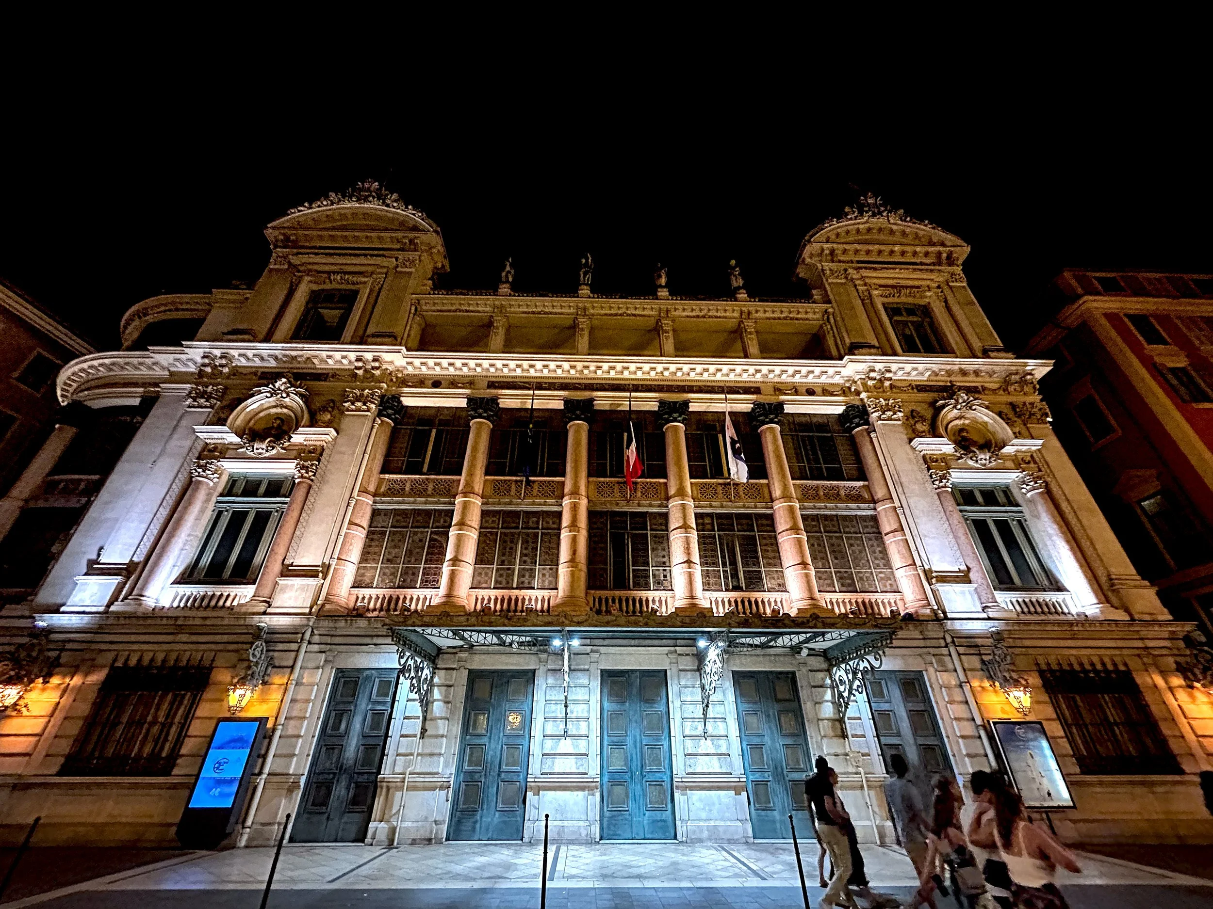 The Opéra de Nice building at night, with a brightly lit, ornate facade and people walking in front