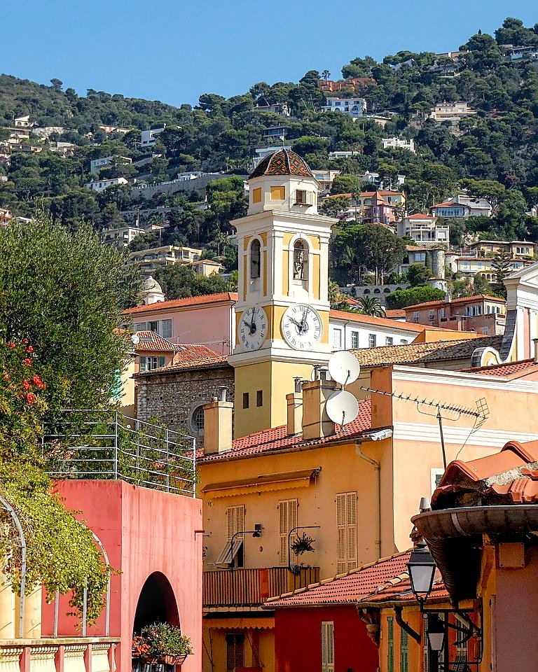 A yellow clock tower with two clocks in the old town of Nice, with a hillside and houses in the background