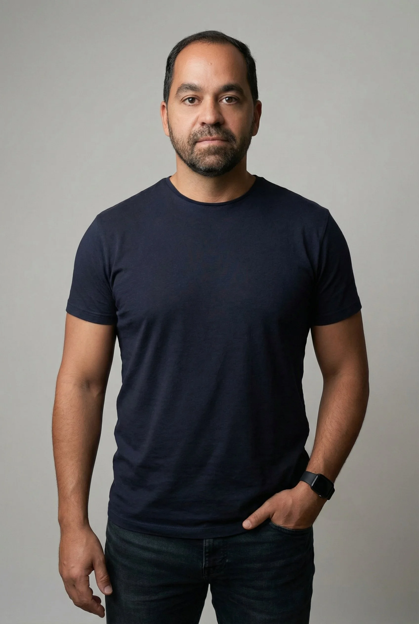 Homem de cabelo escuro, barba por fazer, usando camiseta preta e relógio preto, posando com a mão no bolso esquerdo, contra fundo cinza claro.