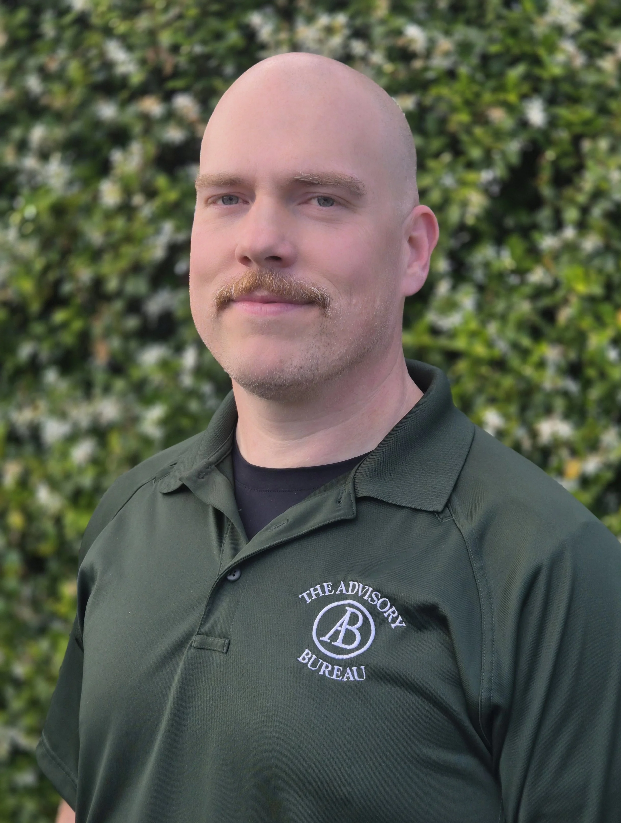 A man with a bald head and mustache wearing a black shirt and backpack against a dark background.