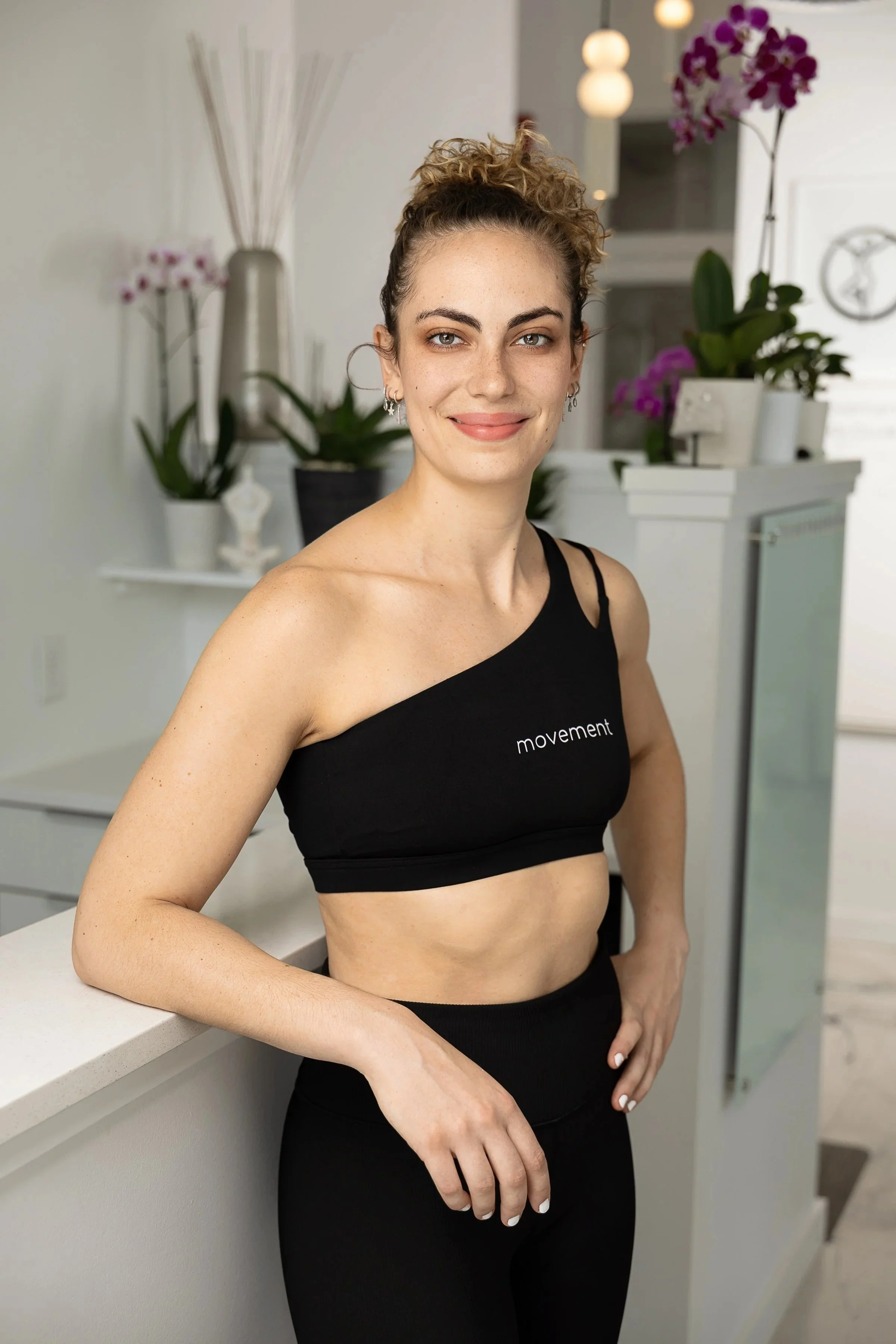 Black and white photo of a young woman with curly hair tied up in a bun, wearing a sleeveless crop top and high-waisted leggings, standing in front of a brick wall and a dark background, smiling at the camera.