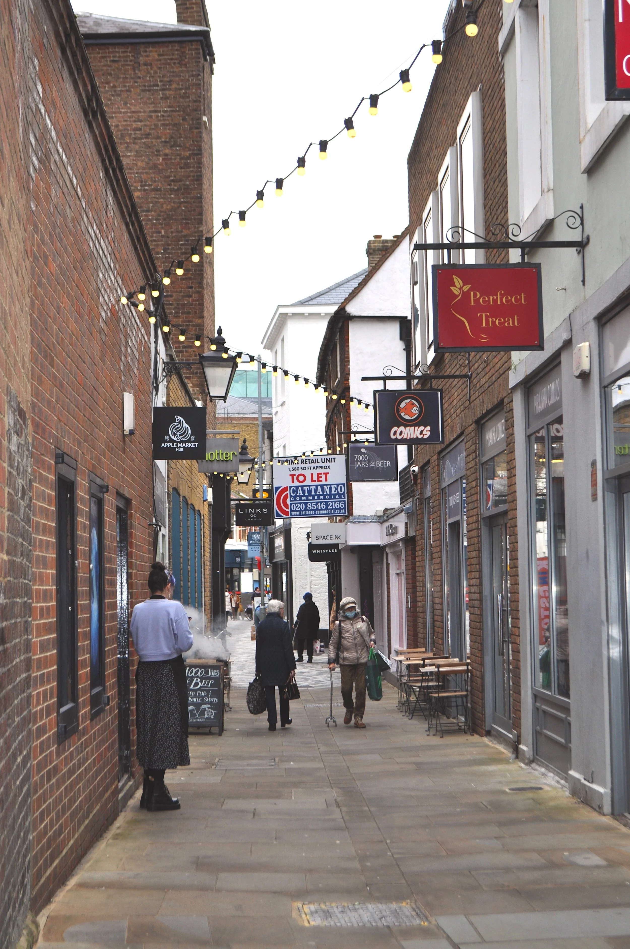 A narrow city street with brick buildings on both sides, string lights hanging overhead, and several pedestrians walking. There are various signs for shops and businesses, some with outdoor seating.