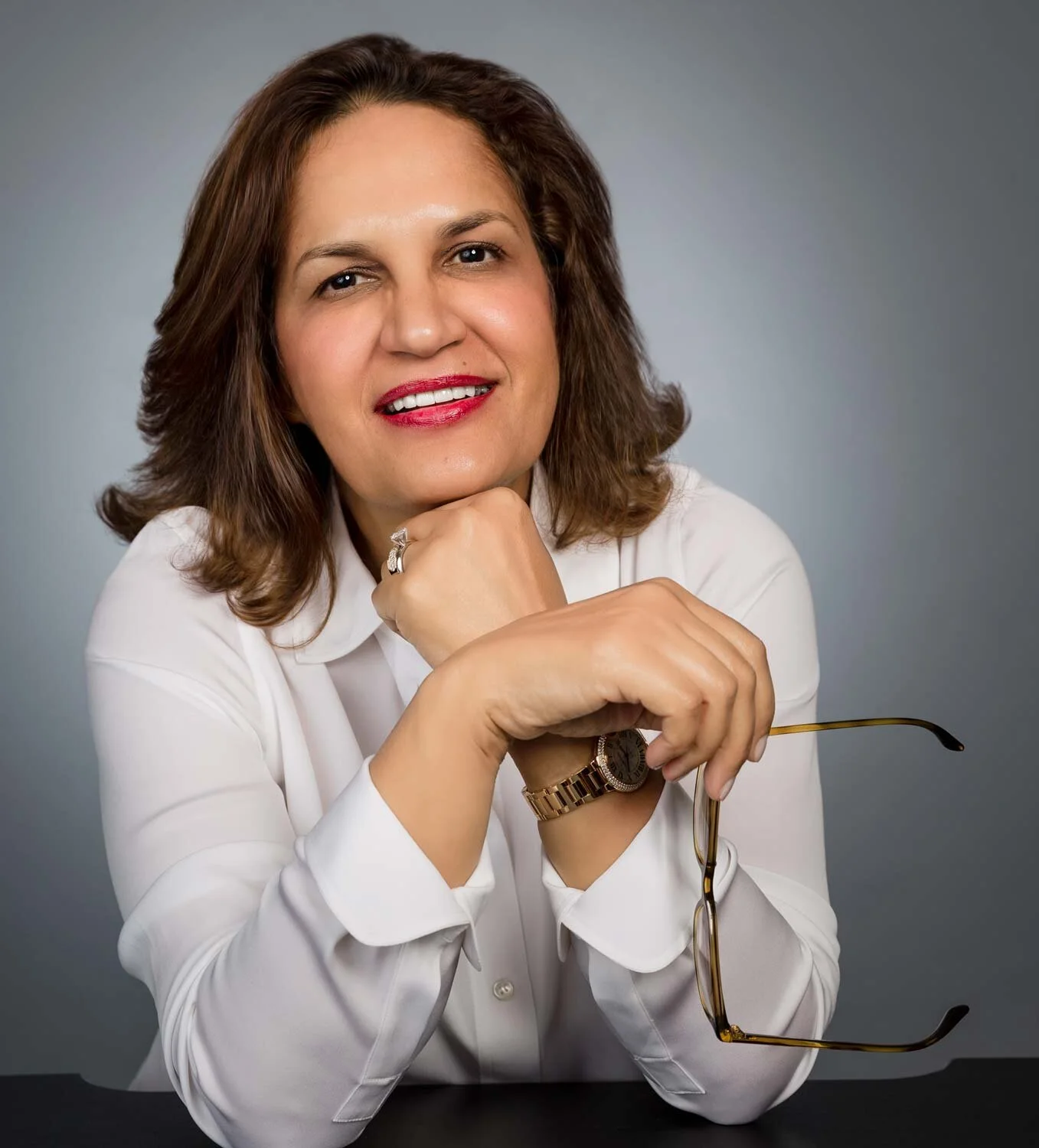 Portrait of a woman with brown hair, wearing a white shirt, red lipstick, a watch, and a ring, holding glasses and smiling at the camera against a grey background.