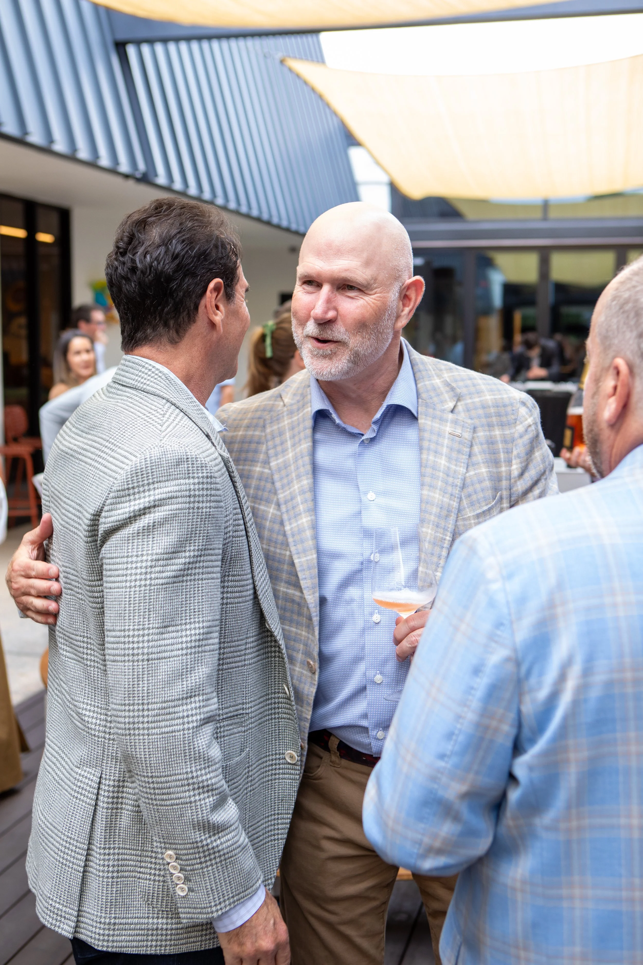 Three men chatting at an outdoor social event, one holding a glass of rosé wine with others dressed in checkered blazers.