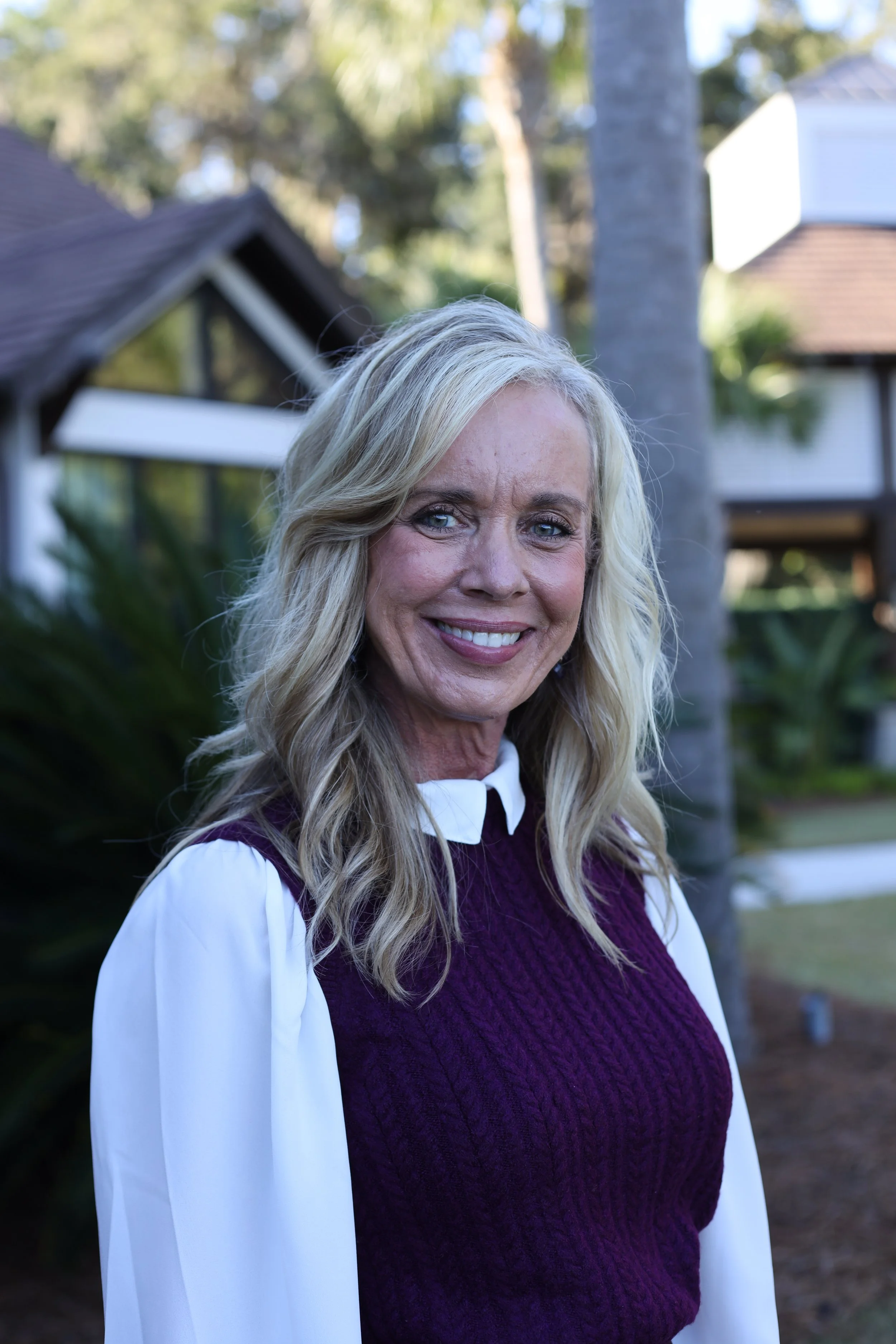 A smiling woman with blonde hair wearing a black blazer and white blouse, standing indoors with a mirror and houseplants in the background.