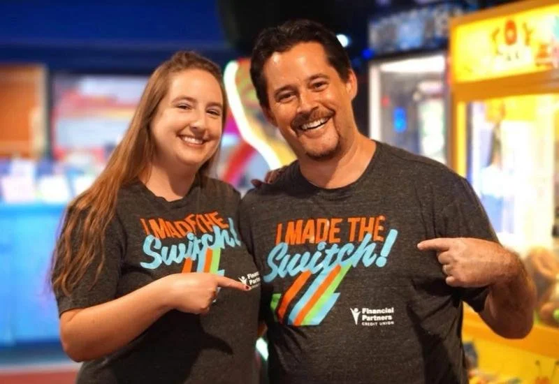 Two people smiling and pointing at their matching T-shirts that say "I MADE THE SWITCH!" in front of an arcade at a bowling alley.