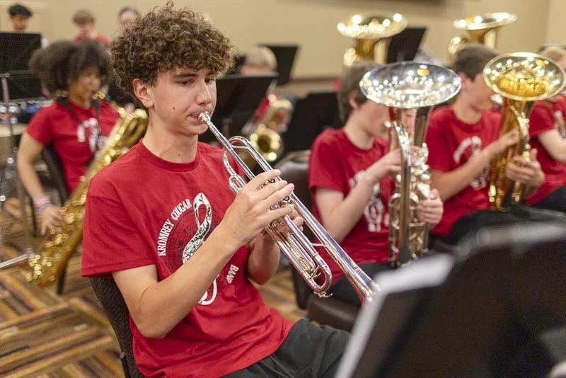 Young boy playing trumpet in band practice, with other students playing various brass instruments in the background, all wearing matching red shirts.