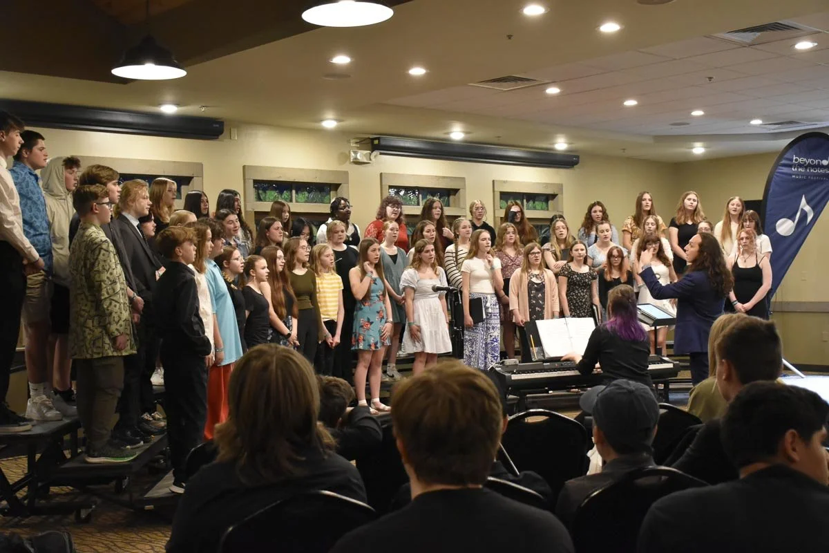 A choir of children and teenagers performing on stage during a concert, with a woman conducting and a pianist accompanying, audience members seated in front.