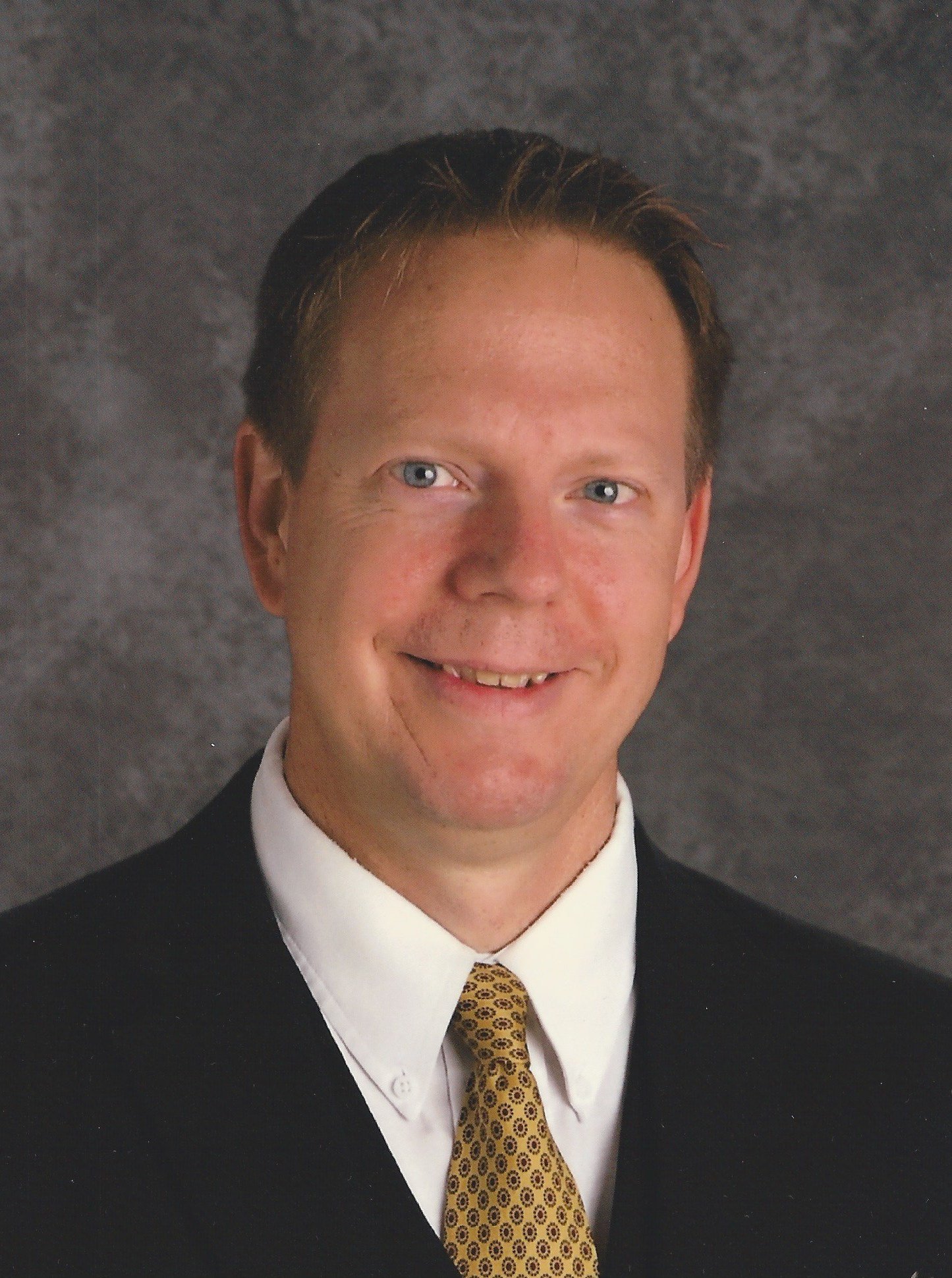 A smiling man with light skin, blue eyes, and short brown hair, dressed in a suit with a white shirt and yellow patterned tie, against a grey textured background.