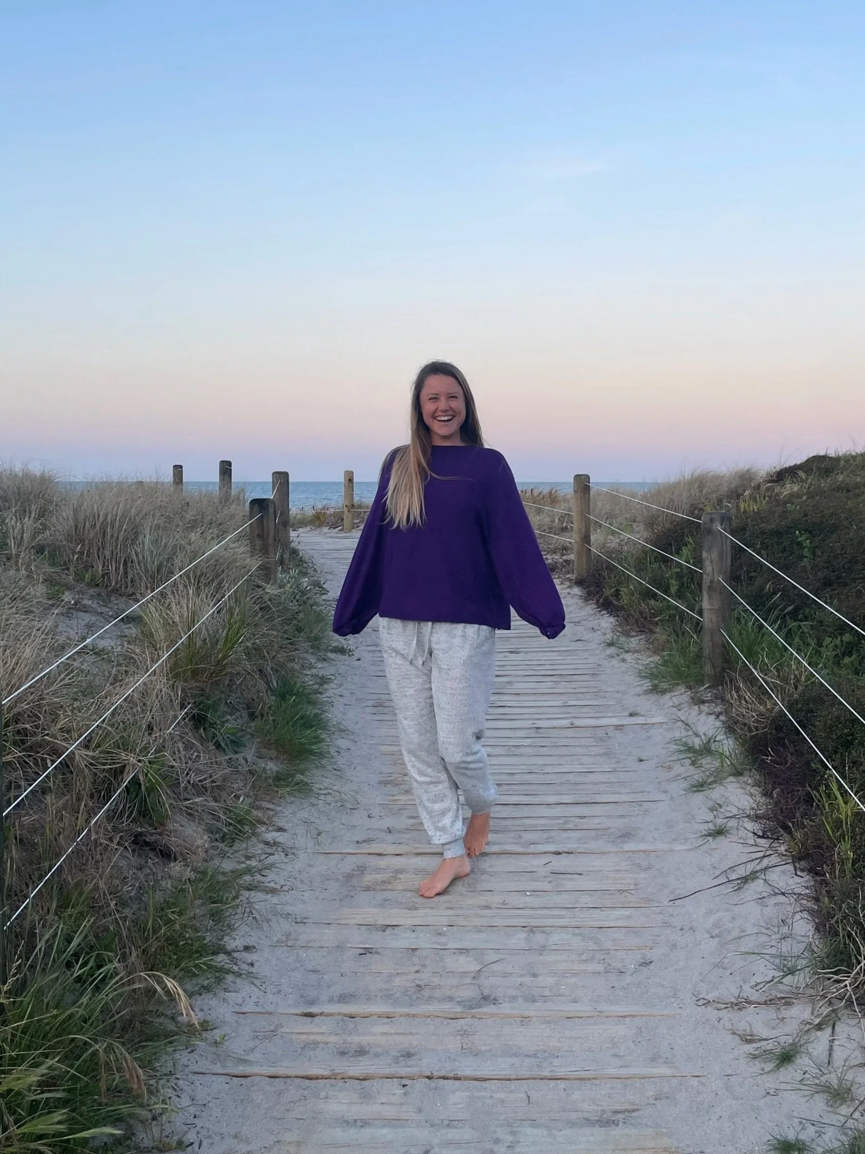 A young woman with long blonde hair smiling and walking barefoot on a wooden pathway through a sandy beach area with grass and bushes, wearing a purple sweatshirt and light gray pants, during sunset or sunrise with calm ocean and pale sky in the background.