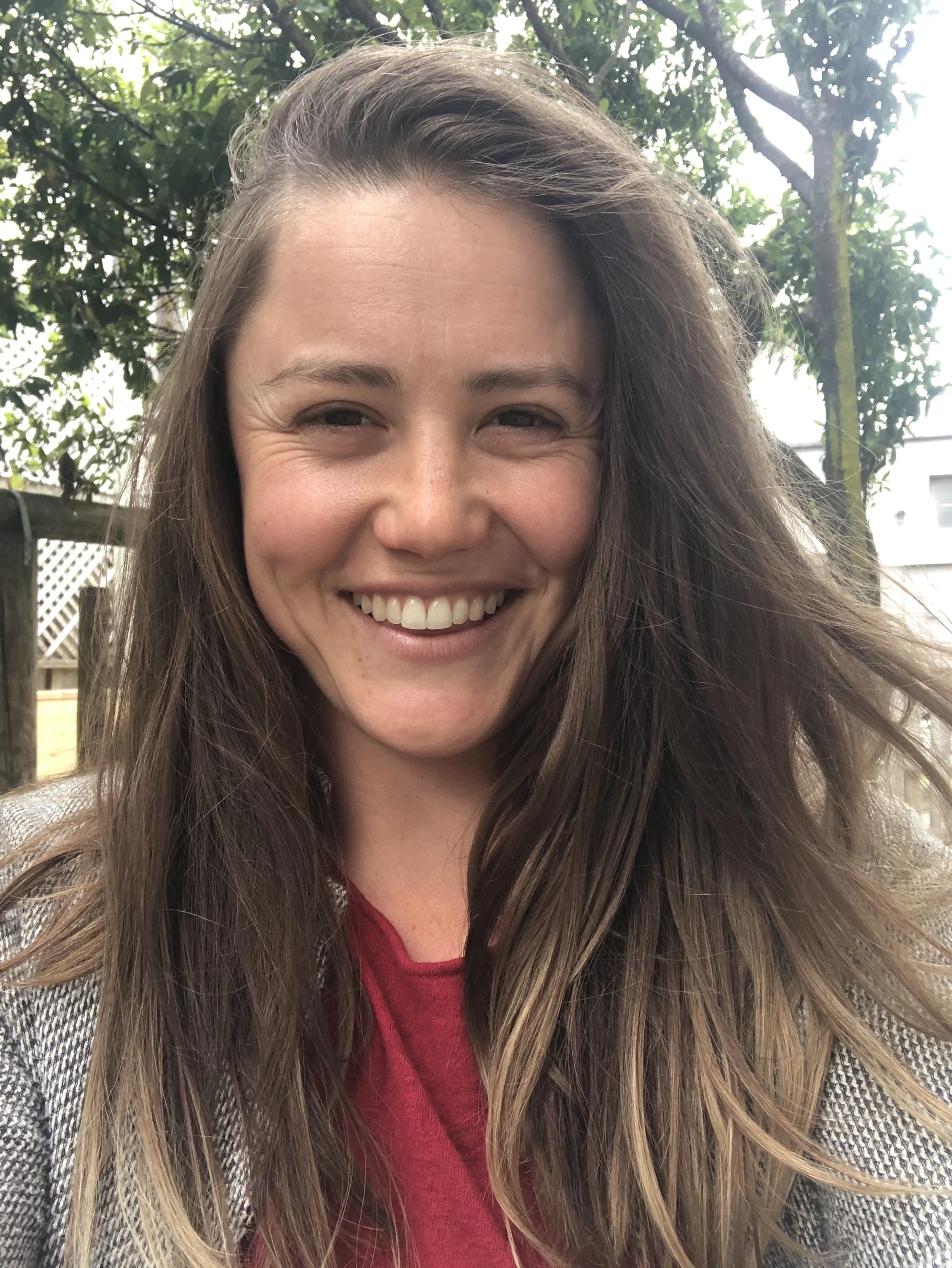 A woman with long brown hair smiling outdoors with greenery and a white building in the background.