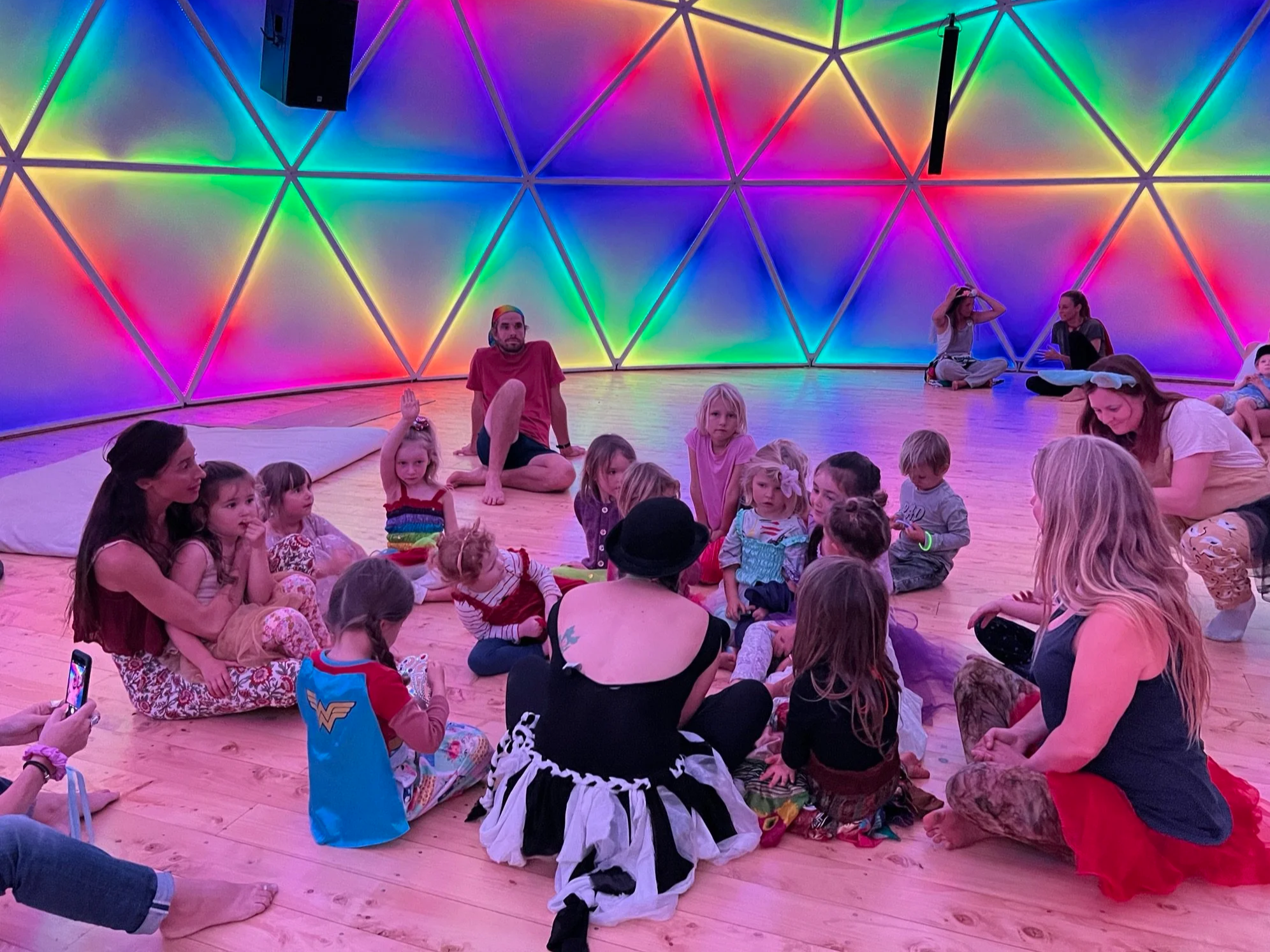 A group of children and adults sitting on a wooden floor inside a geodesic dome with colorful rainbow lighting. Some children are engaged with an adult, and others are sitting quietly. The dome's wall has vibrant, multi-colored lights forming a geometric pattern.