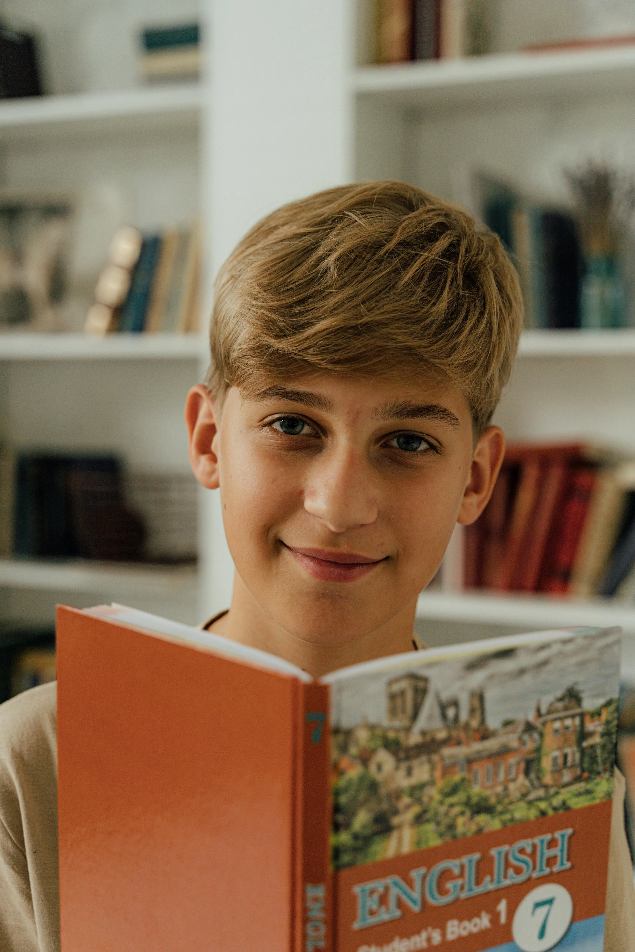 Young boy holding an open English language textbook, smiling in a room with bookshelves in the background.
