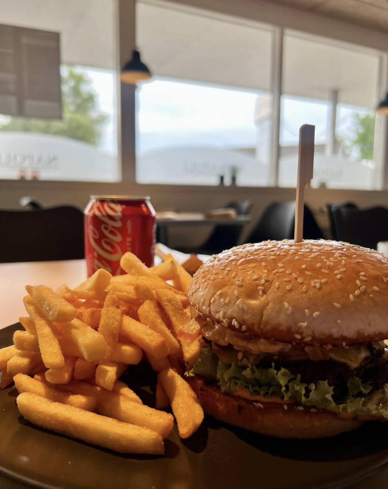 A plate of French fries, a cheeseburger with lettuce and tomato, and a can of Coca-Cola on a table in a restaurant with large windows and patio umbrellas outside.